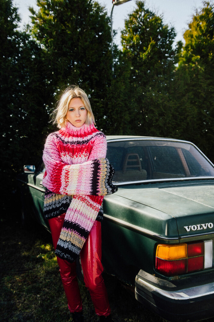 Foto: Pontus Hammarström Artisten Julia Adams i stickad rosa tröja och glittriga byxor framför en gammal Volvo.