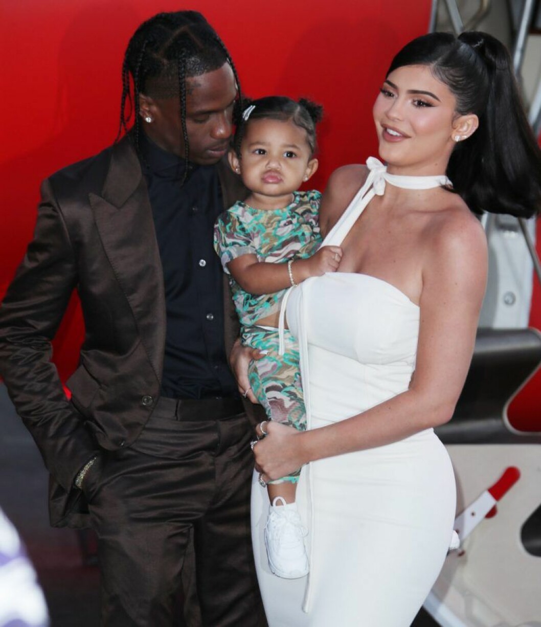 Kylie Jenner står i vit klänning och håller dottern Stormi och Travis Scott är på väg att pussa Stormi på kinden